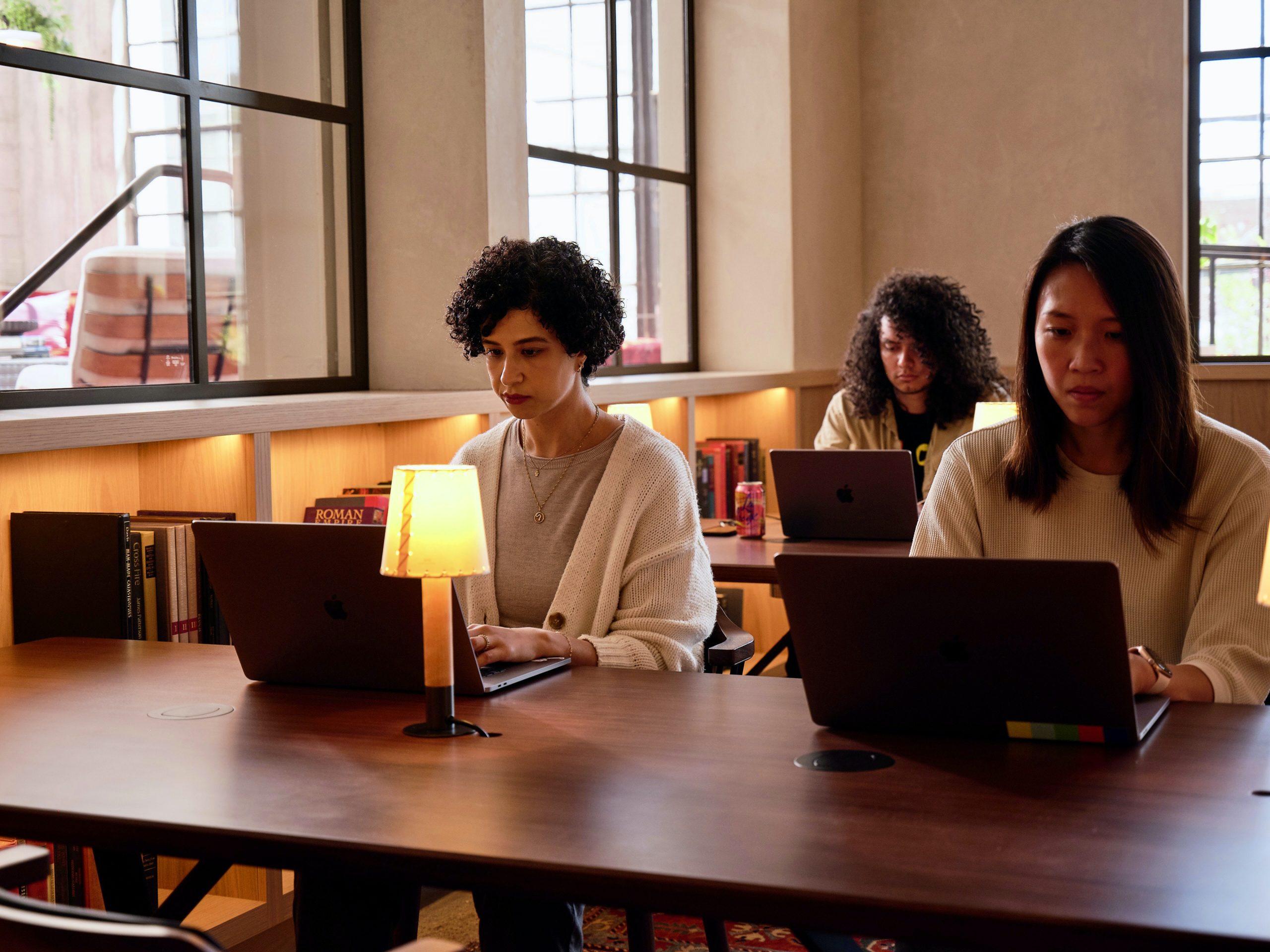 People sitting in a warmly lit library working on laptops at long tables
